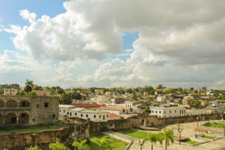 Eine malerische Aussicht auf die Altstadt von Santo Domingo in der Dominikanischen Republik, mit historischen Gebäuden, grünen Palmen und einem bewölkten Himmel.