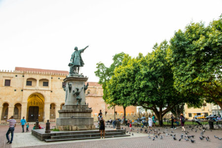 Eine historische Statue von Christoph Kolumbus steht im Zentrum eines lebendigen Platzes in Santo Domingo, umgeben von alten Gebäuden und grünen Bäumen. Menschen spazieren umher und Tauben fliegen in der Nähe.